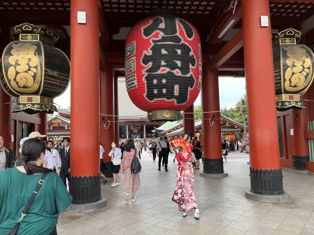 Geisha in traditional kimono posing in front of the Hozomon Gate at Senso-ji Temple, with the iconic red structure and giant lantern in Tokyo, Japan.