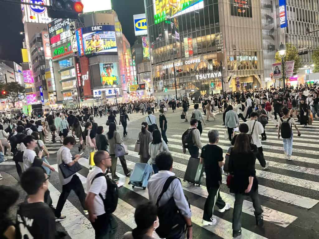 Pedestrians navigate through Tokyo's Shibuya Crossing, with colorful billboards and the famous Hachiko statue in the background.