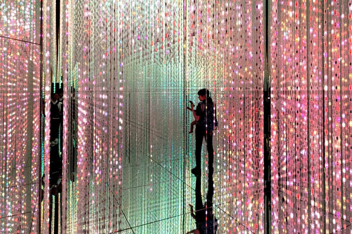 Visitors interacting with the Forest of Resonating Lamps at TeamLab Borderless, featuring multicolored, illuminated lamps hanging from the ceiling.