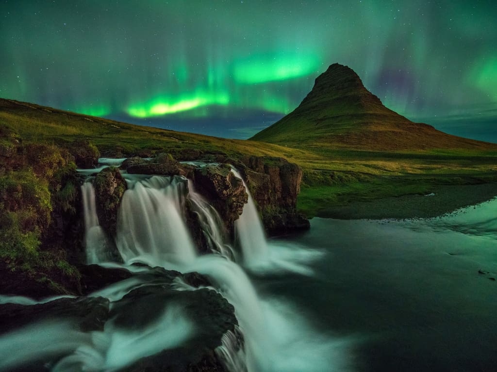 The silhouette of Kirkjufell under the starry sky lit by the green glow of the Aurora Borealis.